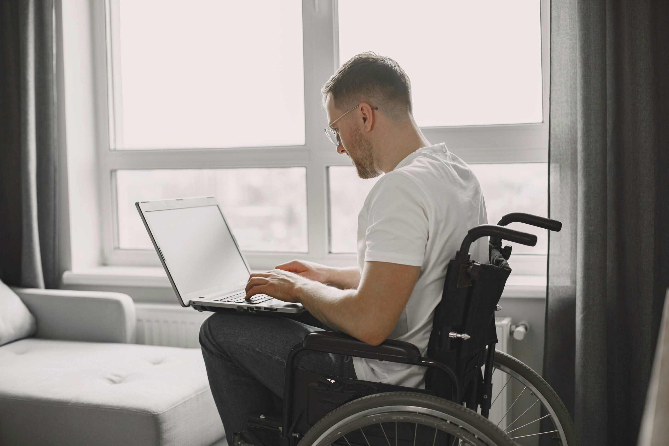 A man in a wheelchair is using a laptop in a bright room with a large window.